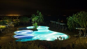 Una piscina iluminada de noche con un diseño singular, rodeada de sillones de descanso y sombrillas.