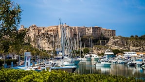 Un puerto deportivo con barcos amarrados, un edificio de varios pisos con arcos, y un acantilado rocoso al fondo.