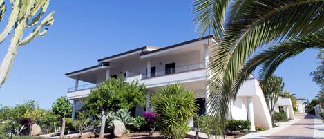 Un edificio de dos pisos con balcones, rodeado de jardines ajardinados y palmeras.