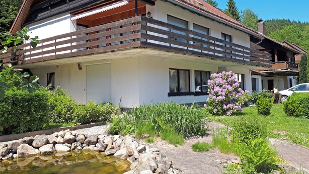 Ein zweistöckiges Haus mit Balkon, umgeben von üppigem Grün und einem kleinen Teich.