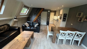 A modern loft apartment with a dining area, a living room with a black leather sofa, and a kitchen in the background.