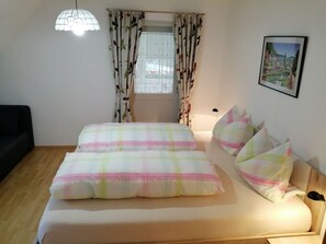 A neatly made bed with a plaid bedspread and pillows, a window with floral curtains, a black chair, and a framed picture on the wall. - Pretty Apartment in Oppenau near Black Forest National Park (Oppenau)