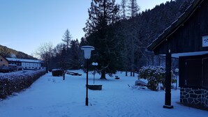A snow-covered outdoor area with a wooden building, a stone wall, and a snow-covered tree. - Wohnung in Nahe Harzwald (Clausthal-Zellerfeld)