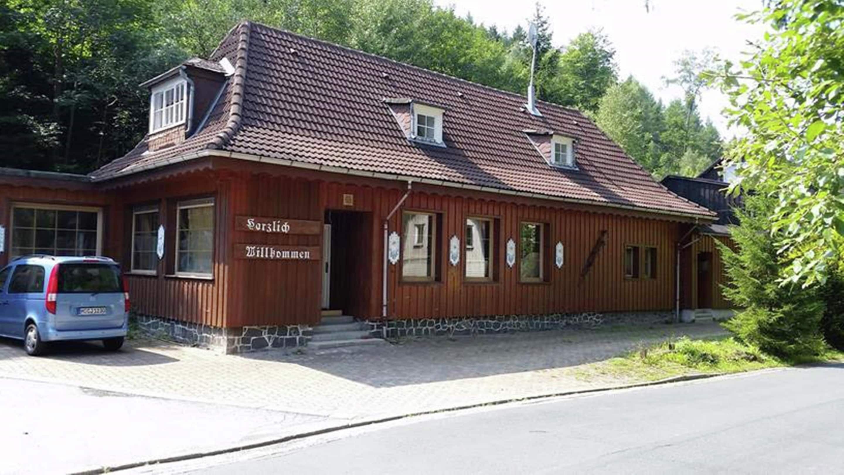 A wooden building with a tiled roof, a sign reading 'Willkommen,' and a parked car.