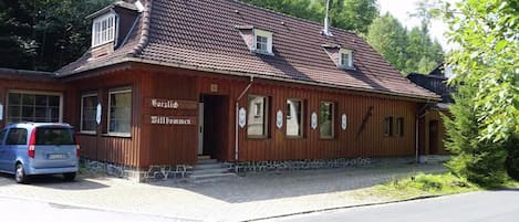 A wooden building with a tiled roof, a sign reading 'Willkommen,' and a parked car.