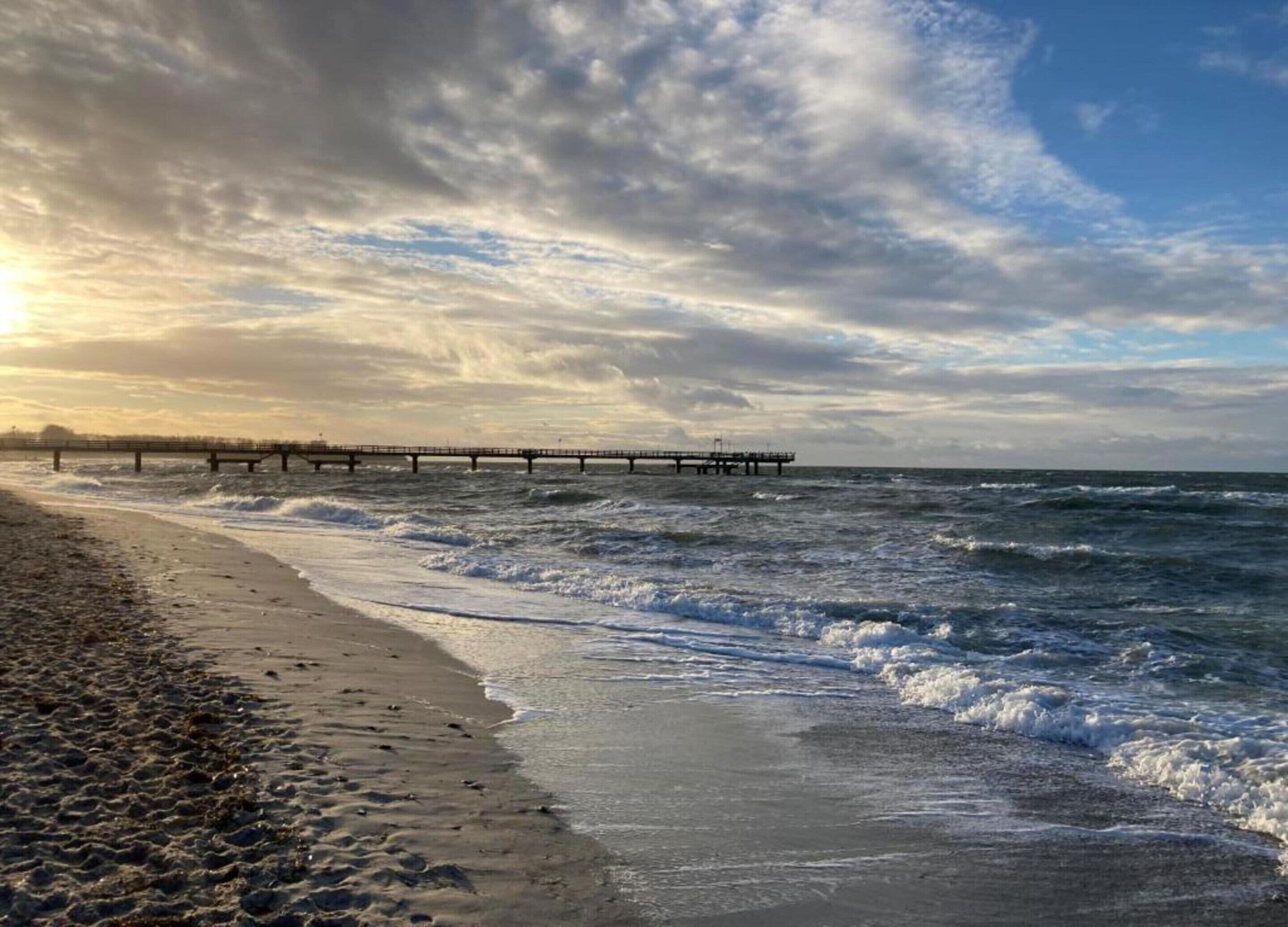 A beach with a pier extending into the ocean, waves crashing onto the shore, and a partly cloudy sky.