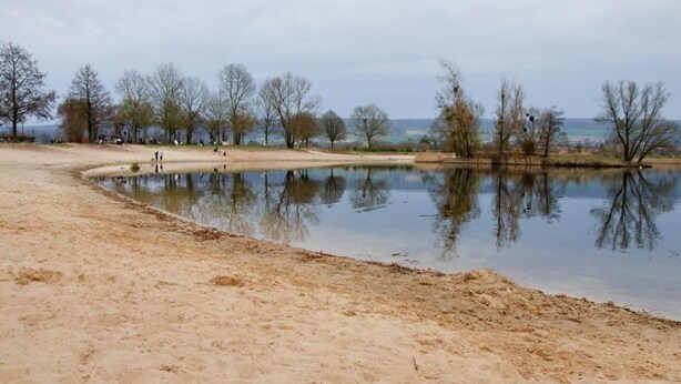 A sandy beach area with a calm water body, trees, and a distant view of a town. - Wohnung in Hoxter Nahe Schloss Corvey (Hoexter)