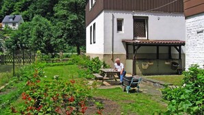 Una casa con jardín, una persona sentada en un banco y un carretilla.