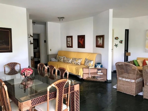 A living room with a glass dining table, wicker chairs, a yellow sofa, and a coffee table.