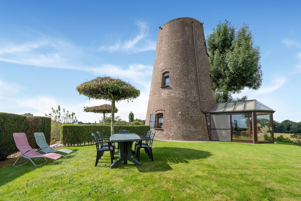 A brick tower with a glass conservatory, a lawn with chairs, and a hedge.