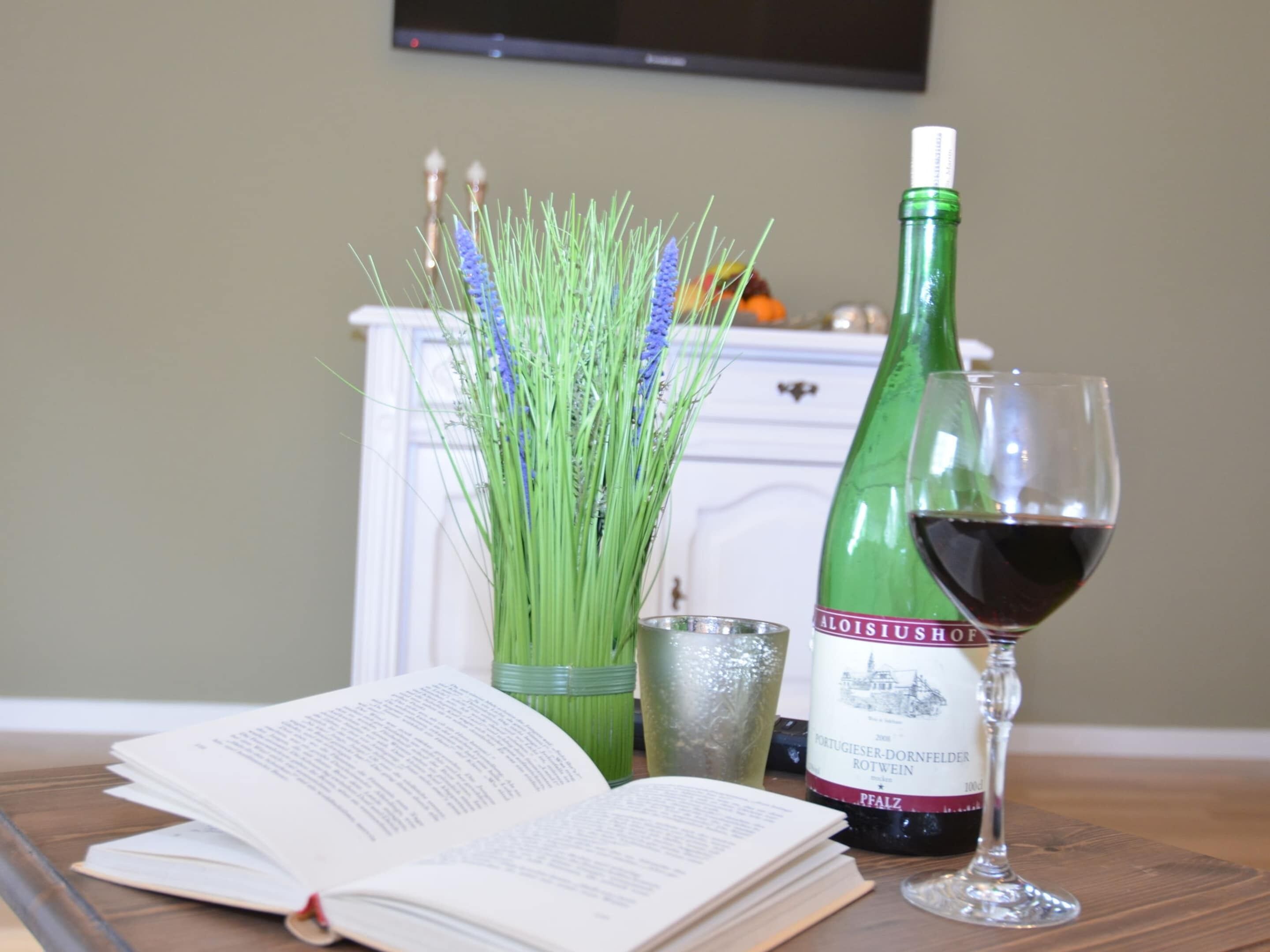 Une bouteille de vin, un verre de vin, un livre et un vase avec des plantes vertes sur une table en bois.