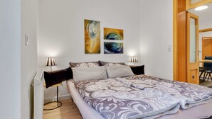 A bedroom with a bed, two bedside lamps, a radiator, and three abstract paintings on the wall.