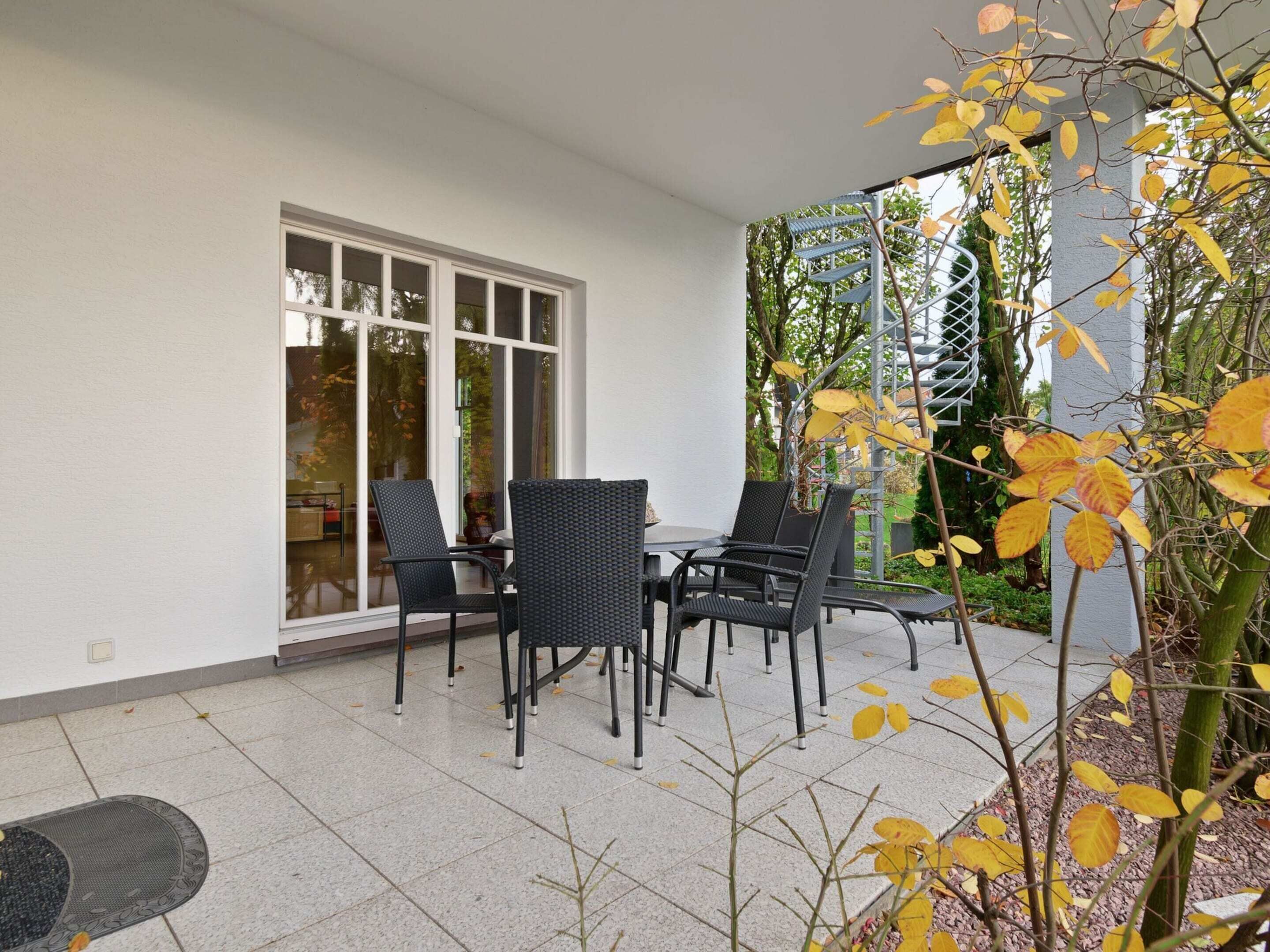 Un patio al aire libre con una mesa y sillas, una zona cubierta con una escalera espiral metálica y vistas a árboles y follaje.