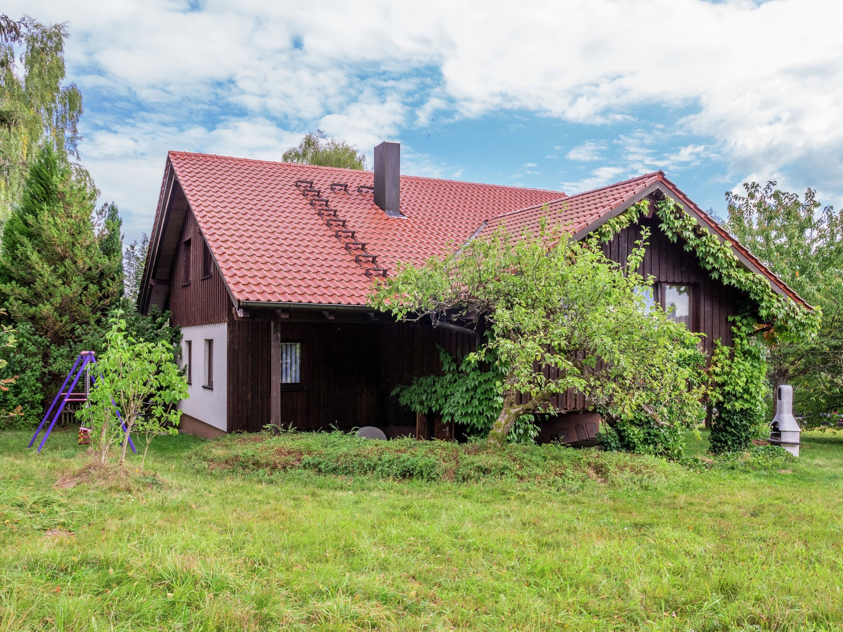 Uma casa de madeira rústica com telhado de telhas vermelhas, rodeada por vegetação e árvores.