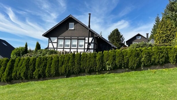 Une maison à deux étages, avec un toit sombre et des encadrements de fenêtres blancs, entourée d’une haie bien entretenue et d’une pelouse verdoyante.