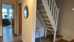 Un escalier avec des rampes blanches menant à l’étage supérieur, une porte avec un miroir, et une vue sur une pièce contenant une chaise et une table.