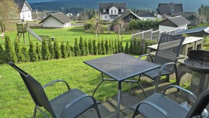 Une terrasse avec une table et des chaises donnant sur un espace verdoyant, avec des maisons et, au loin, une chaîne de montagnes.