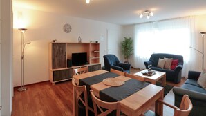Un salon moderne avec une table à manger en bois, deux chaises, un canapé et un téléviseur posé sur un meuble en bois.