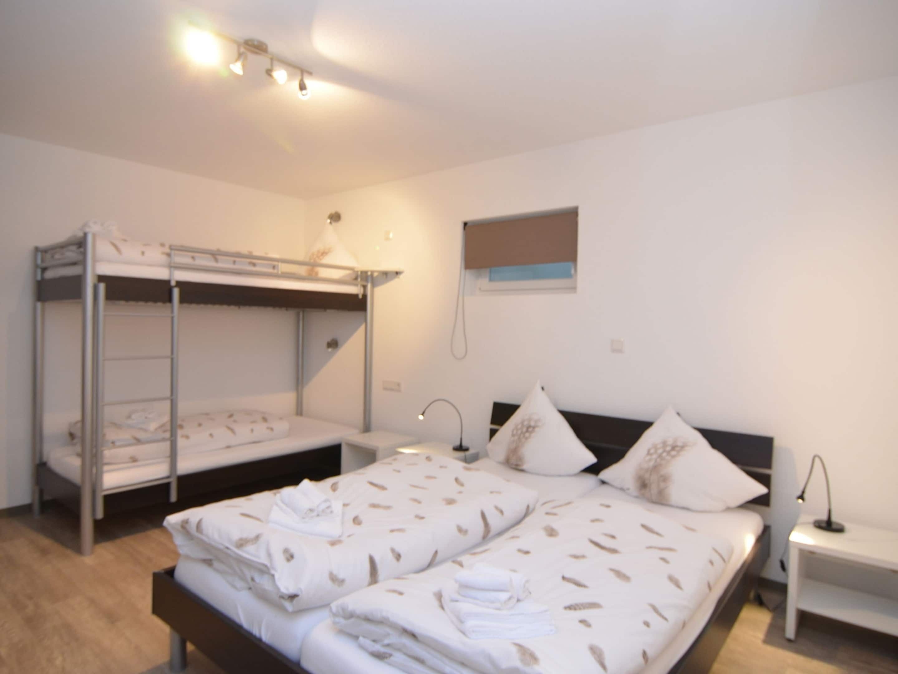 Una habitación con literas, una cama individual, una mesita de noche con lámpara y un pequeño ventanal con persianas.