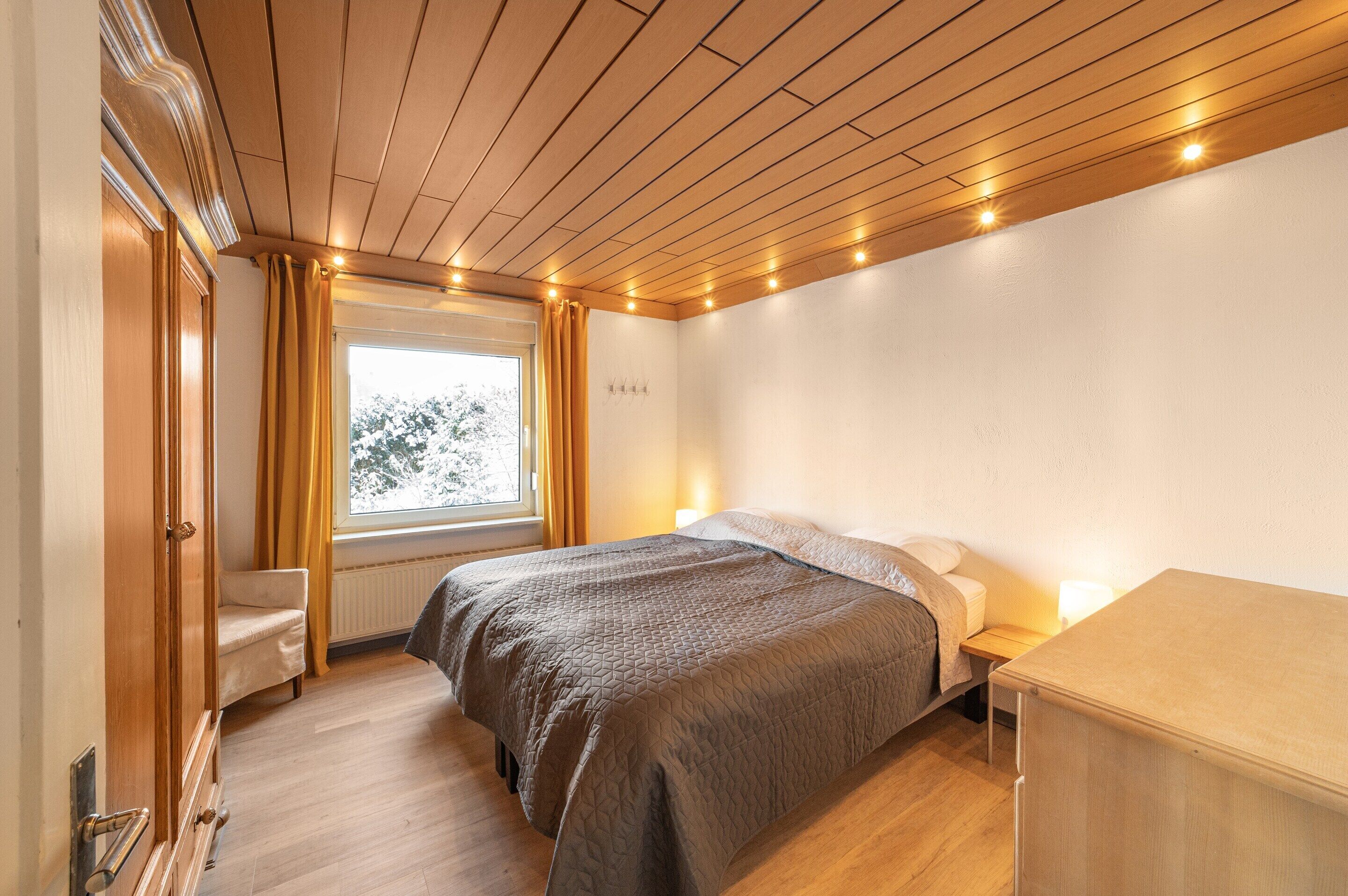 Un dormitorio con techo de madera, una cama con colcha acolchada, una mesita de noche con lámpara y una ventana con cortinas.