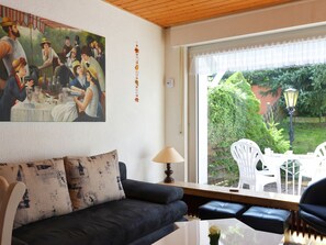 Un salon avec un canapé aux couleurs sombres, une table basse blanche et un tableau représentant une scène de rassemblement social. Une fenêtre offre une vue sur la verdure et sur du mobilier d’extérieur.