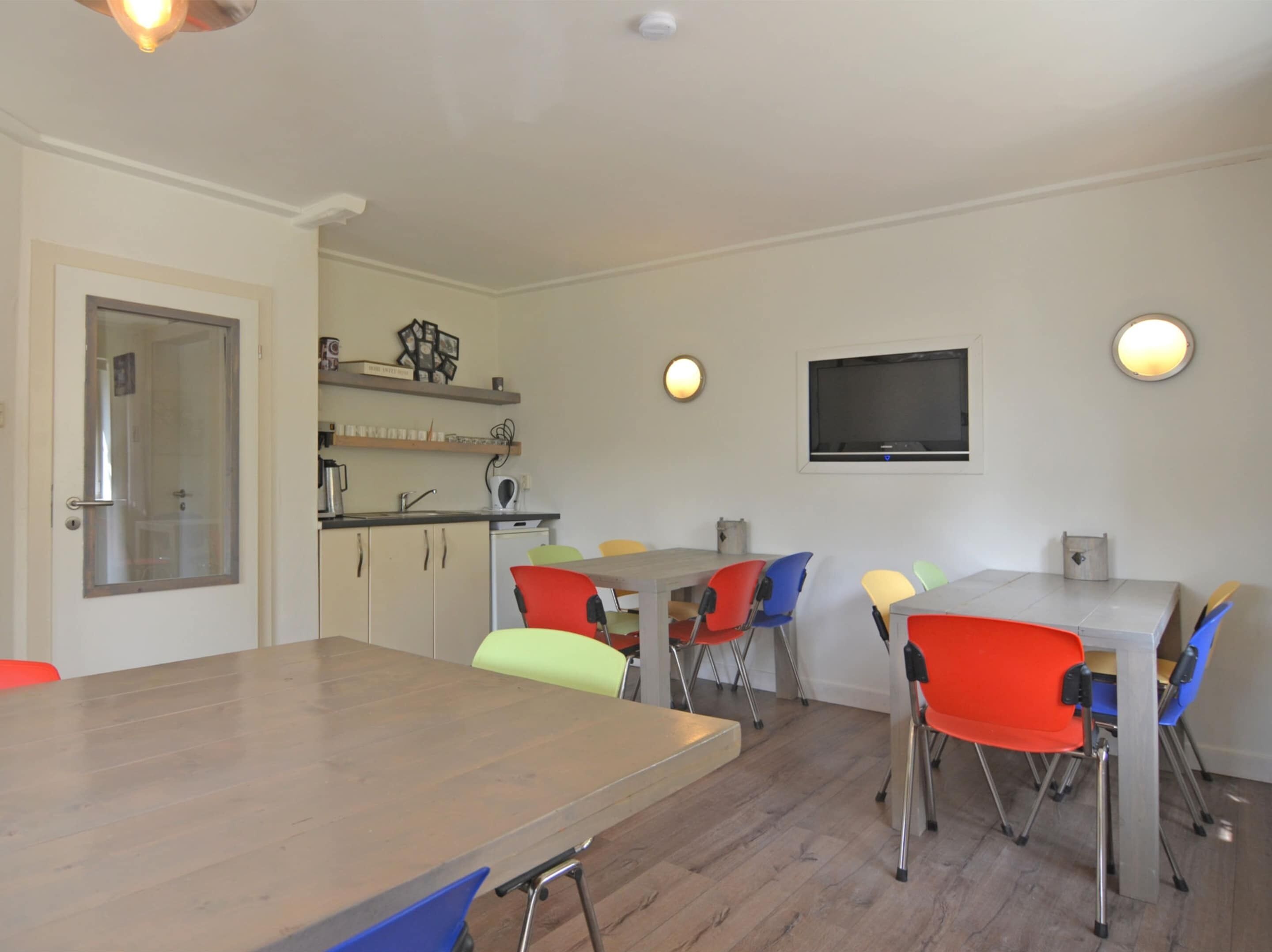 Un comedor con una mesa de madera y sillas de colores, una cocina con un lavaplatos y armarios, y un televisor montado en la pared.