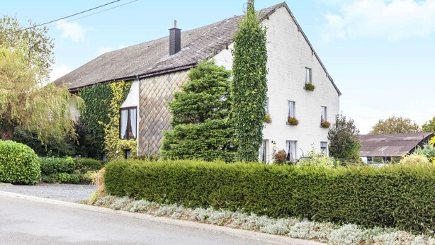 A two-story house with a white exterior, a tiled roof, and a chimney. There are windows with curtains, and the house is surrounded by well-maintained hedges and a gravel driveway. - Holiday Home in Bastogne With Garden (Bastogne)