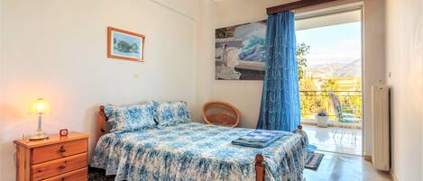 A bedroom with a bed, a wooden nightstand, a lamp, a chair, a painting, and a view of the outdoors.