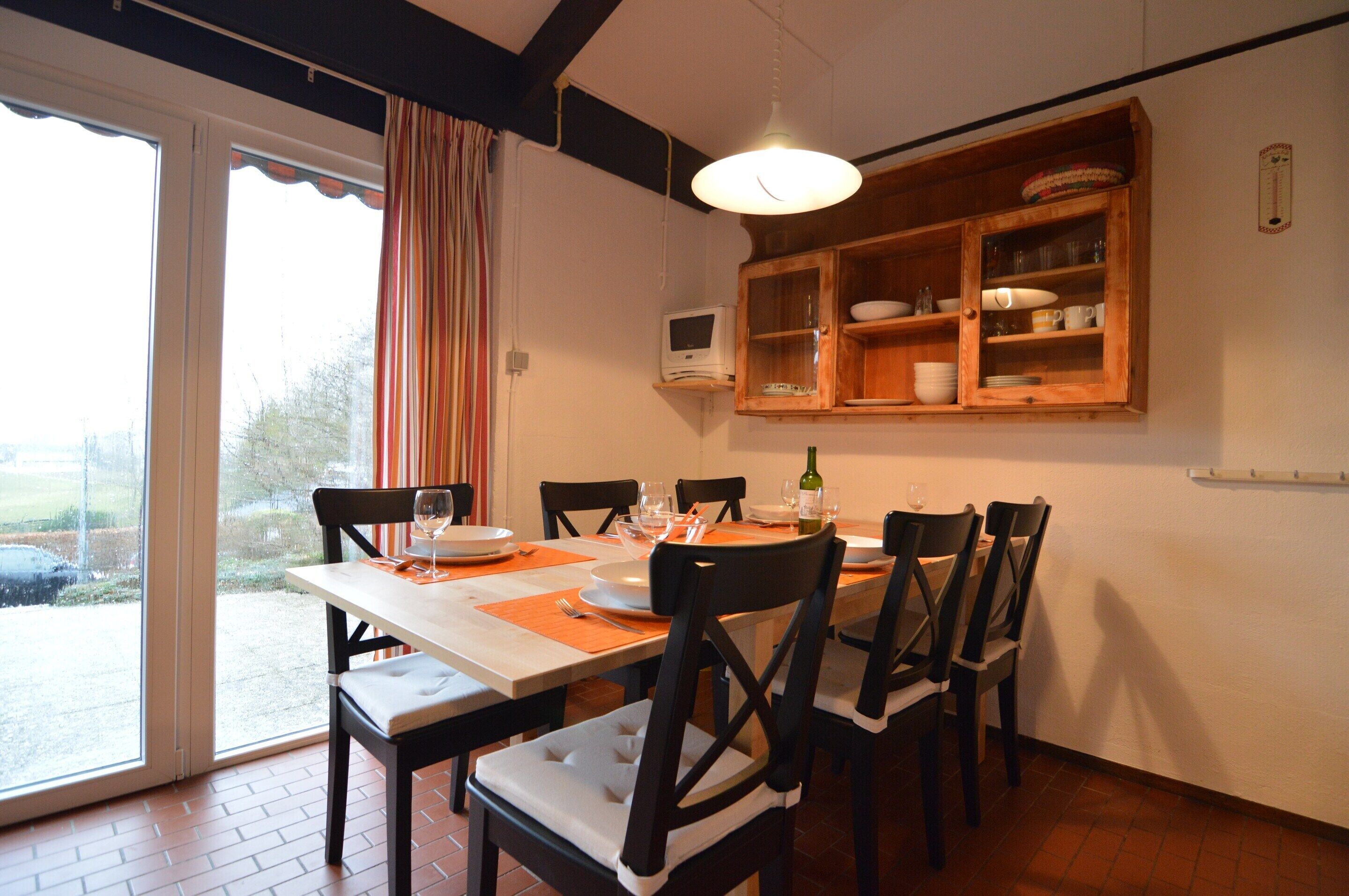 Un comedor con una mesa dispuesta para cuatro, sillas, una ventana con cortinas, un mueble de madera con vajilla, y una lámpara colgante.