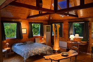 Ein Interieur einer Holzhütte mit einem Bett, einem Esstisch, einem Stuhl, einer Lampe und einem Fenster mit Vorhängen.