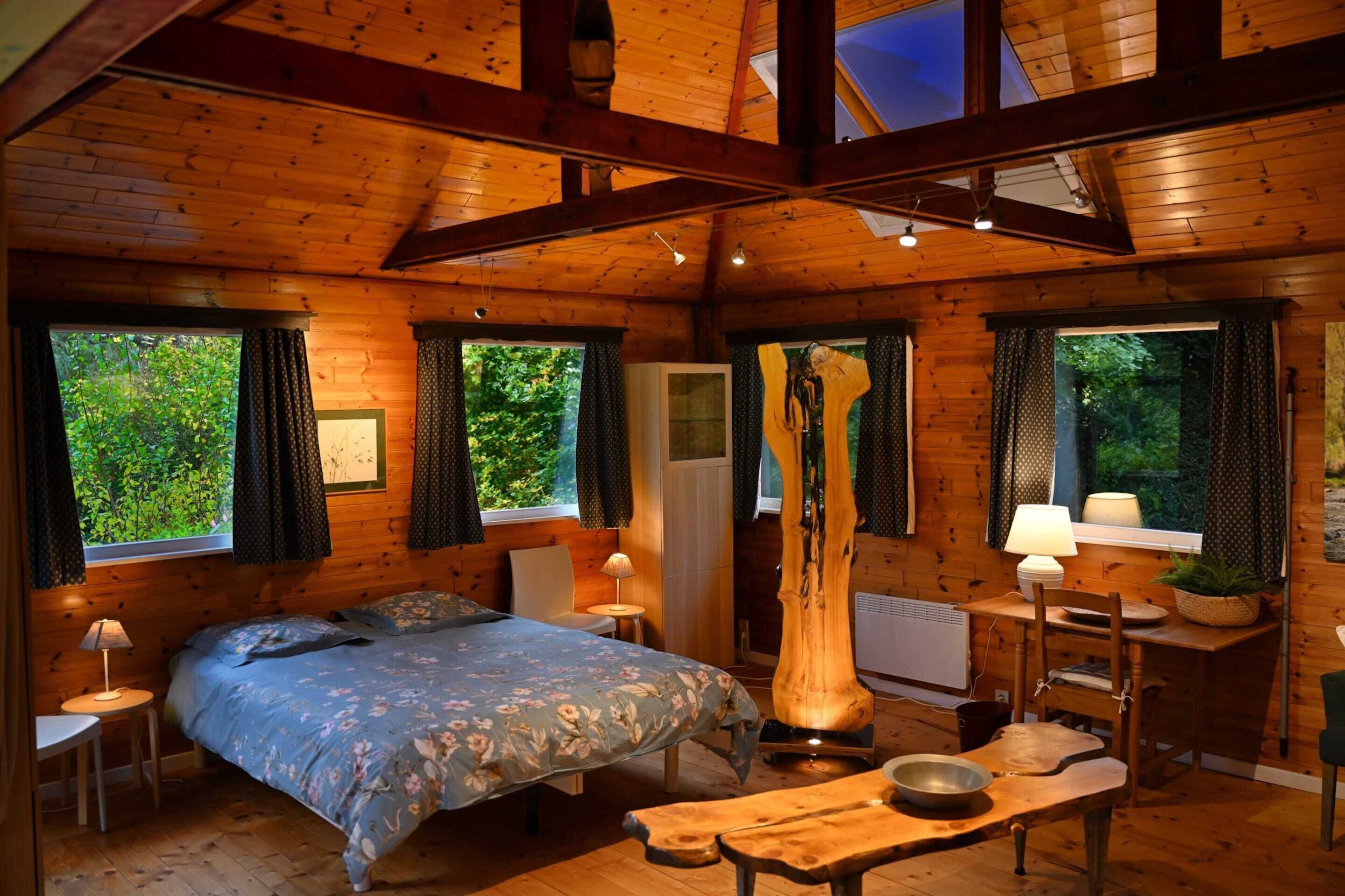 L’intérieur d’un chalet en bois comprend un lit, une table à manger, une chaise, une lampe et une fenêtre avec des rideaux.