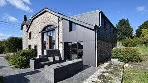 Une maison moderne avec une façade en pierre, de grandes fenêtres et un chemin en pierre menant à un espace aménagé pour s’asseoir.