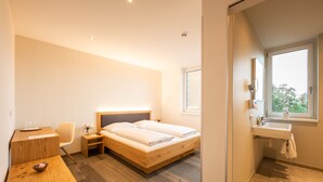Una habitación de hotel moderna con una cama de madera, un escritorio con silla, una mesita de noche, un ventanal con vistas y un baño con lavamanos y espejo.