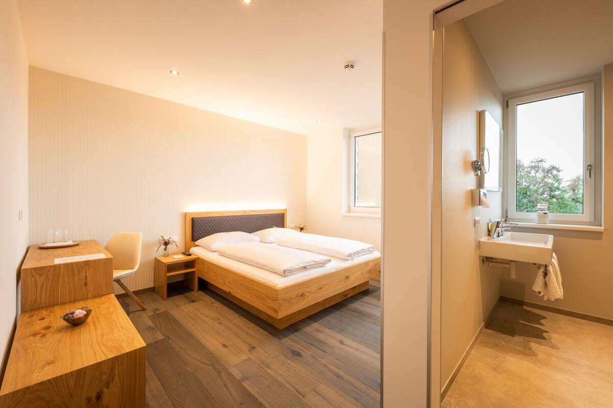 Una habitación de hotel moderna con una cama de madera, un escritorio con silla, una mesita de noche, un ventanal con vistas y un baño con lavamanos y espejo.