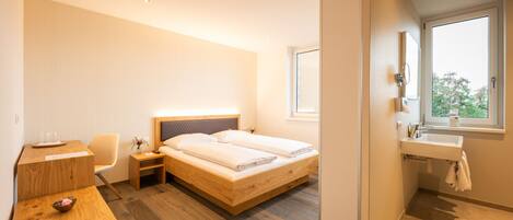 Une chambre d’hôtel moderne avec un lit en bois, un bureau avec une chaise, une table de chevet, une fenêtre donnant sur un paysage agréable et une salle de bain équipée d’un lavabo et d’un miroir.