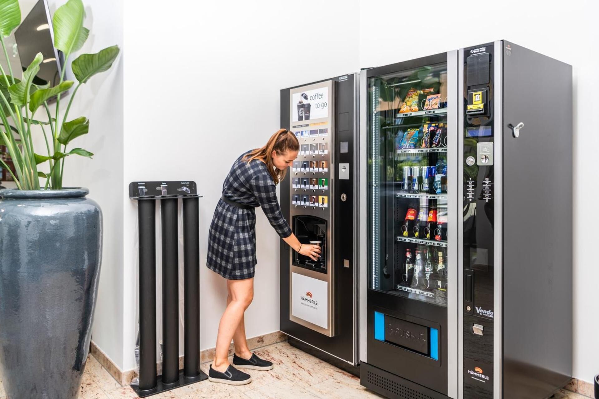 A vending machine with a variety of beverages and snacks, a person interacting with it, and a large potted plant beside it.