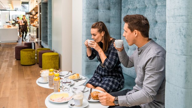 Una pareja desayunando en una mesa, en un interior moderno con pared acolchada y zona de estar.
