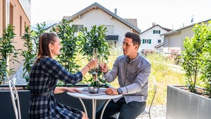 Dos personas sentadas en una mesa al aire libre con una botella de champán, rodeadas de vegetación y edificios residenciales.