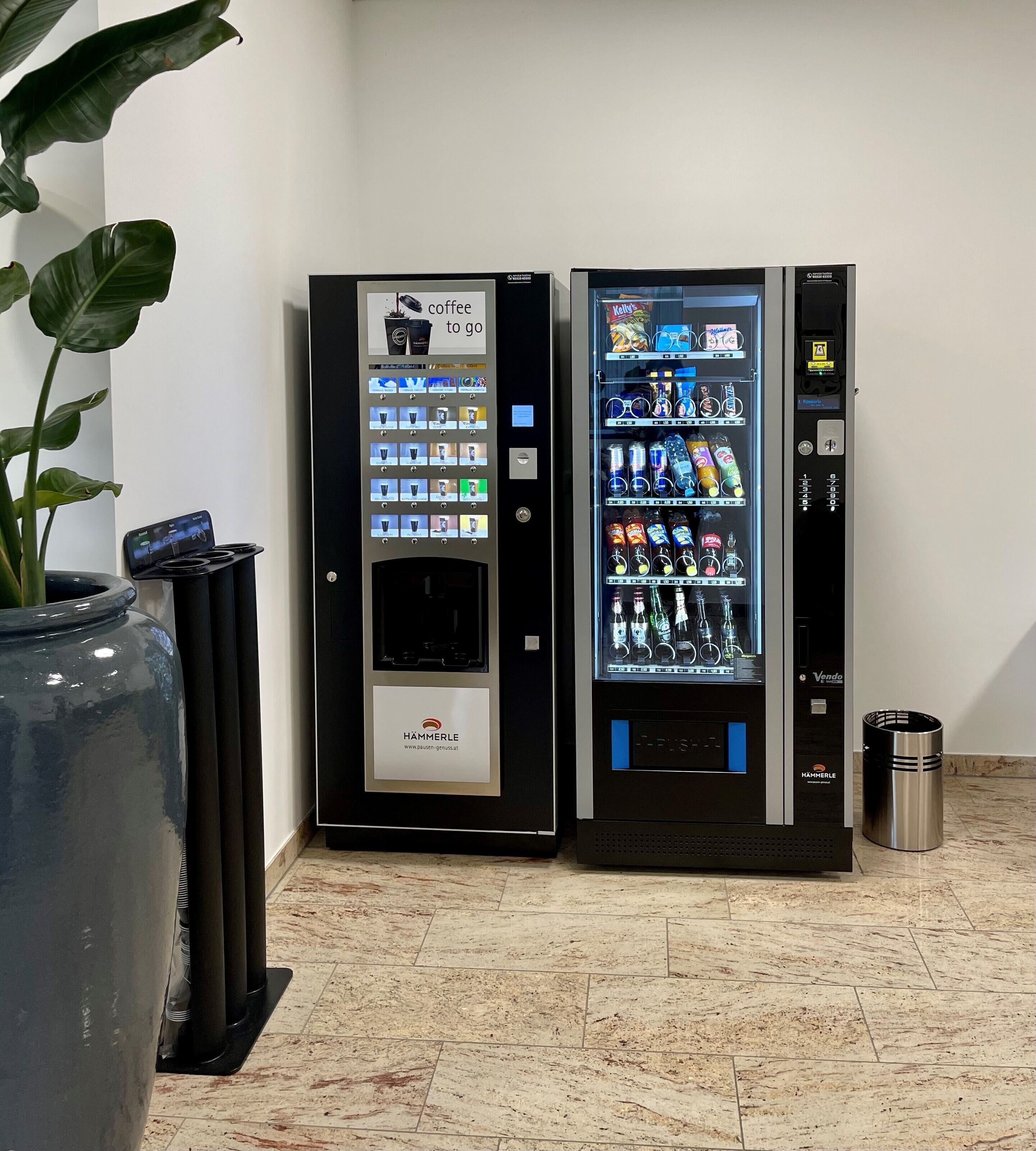 Dos máquinas expendedoras con diferentes bebidas y aperitivos, un cubo de basura y una planta en maceta.