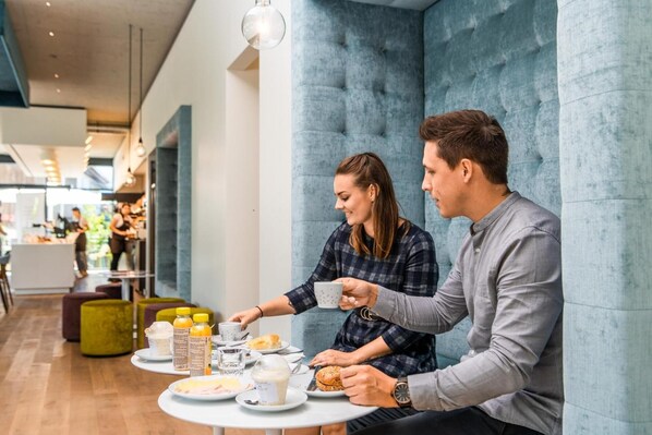 A couple dining at a table with breakfast items, in a modern interior with blue walls and a visible café in the background. - Motel Keckeis Inn (Sulz)
