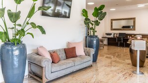 Un vestíbulo moderno con un sofá gris, plantas en macetas grandes, un mostrador de recepción y un espejo.