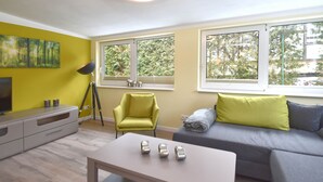 A modern living room with a sofa, armchair, and a coffee table. There are windows with a view of trees and a ceiling light. - Apartment With Private Terrace (Harzgerode)