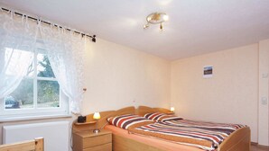 A bedroom with a wooden bed, a bedside table with a lamp, a window with curtains, and a wall-mounted light fixture. - Quaint Holiday Home in Güntersberge near Lake (Harzgerode)