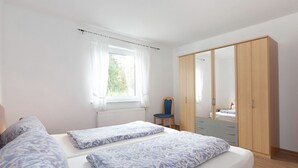 Un dormitorio con cama, una silla, un armario y una ventana con cortinas.