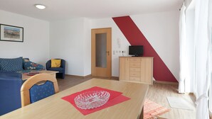 Una sala moderna con una mesa de comedor de madera, un sofá azul, un televisor sobre un mueble bajo y un ventanal con cortinas blancas.