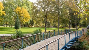 Un parque con un puente peatonal, zonas verdes y árboles con follaje otoñal.