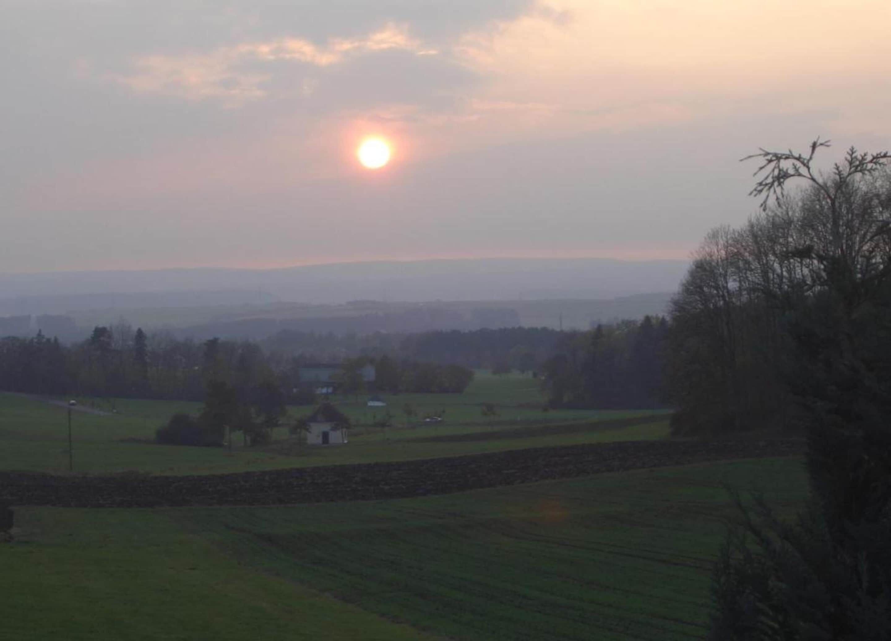 A rural landscape with a clear sky, a setting sun, and a small house surrounded by fields.