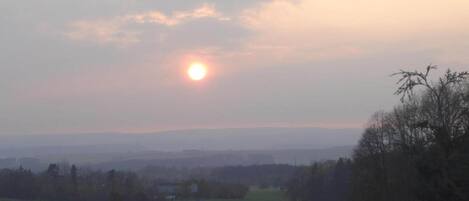 A rural landscape with a clear sky, a setting sun, and a small house surrounded by fields.
