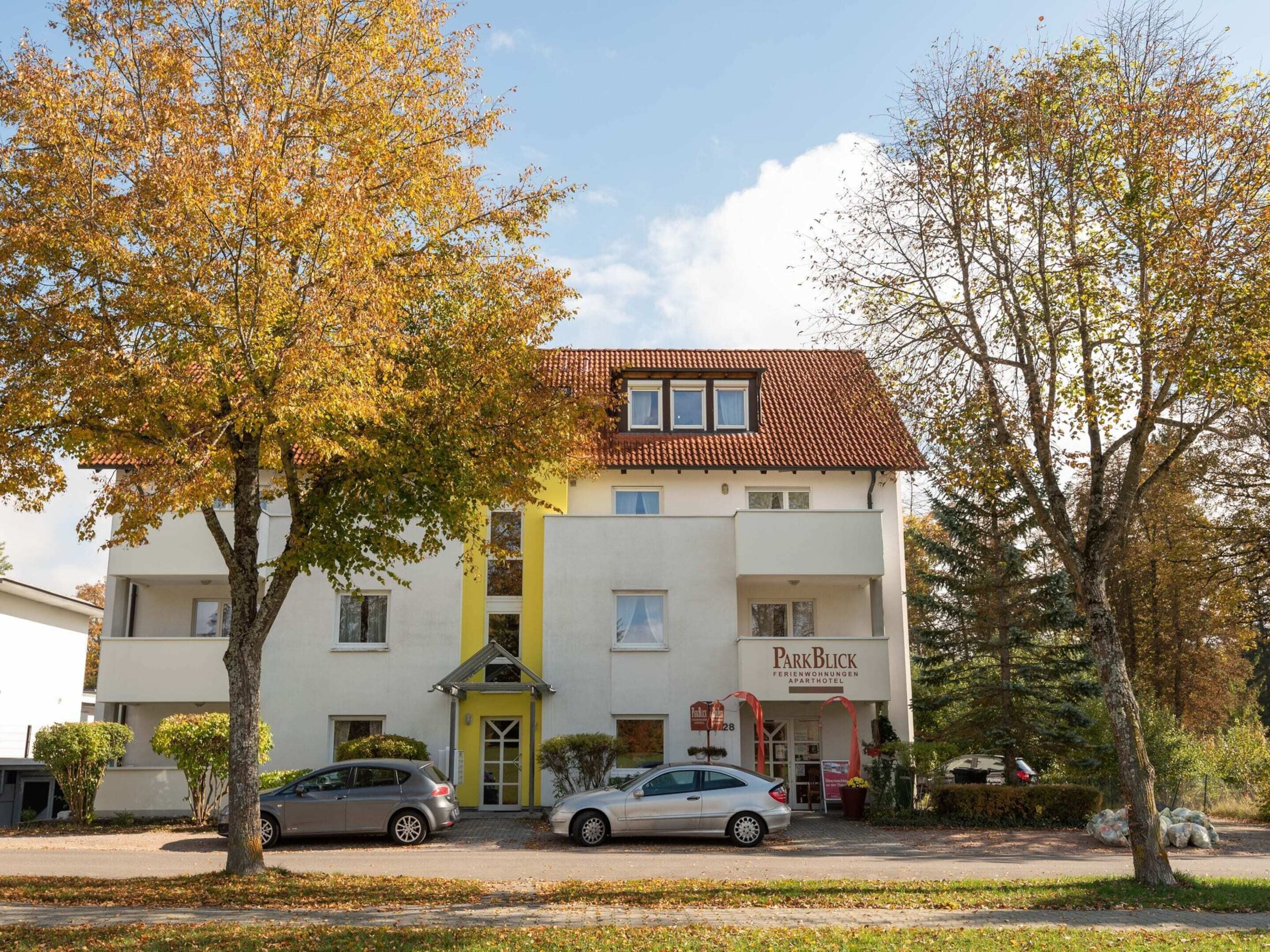 A two-story building with a yellow entrance and a sign reading "ParkBlick Ferienwohnungen Aparthotel".