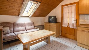 A cozy room with a wooden ceiling, a sofa, a small table, a television, and a kitchen area with cabinets and a window with curtains. - Retreat in the Ammergau Alps (Unterammergau)